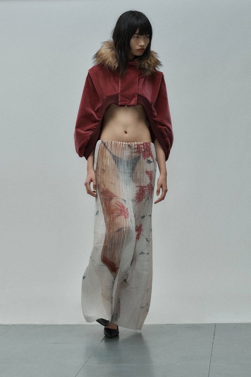 Fashion runway show with a model wearing a velvet jacket with a fur collar and a pleated printed skirt against a grey background.