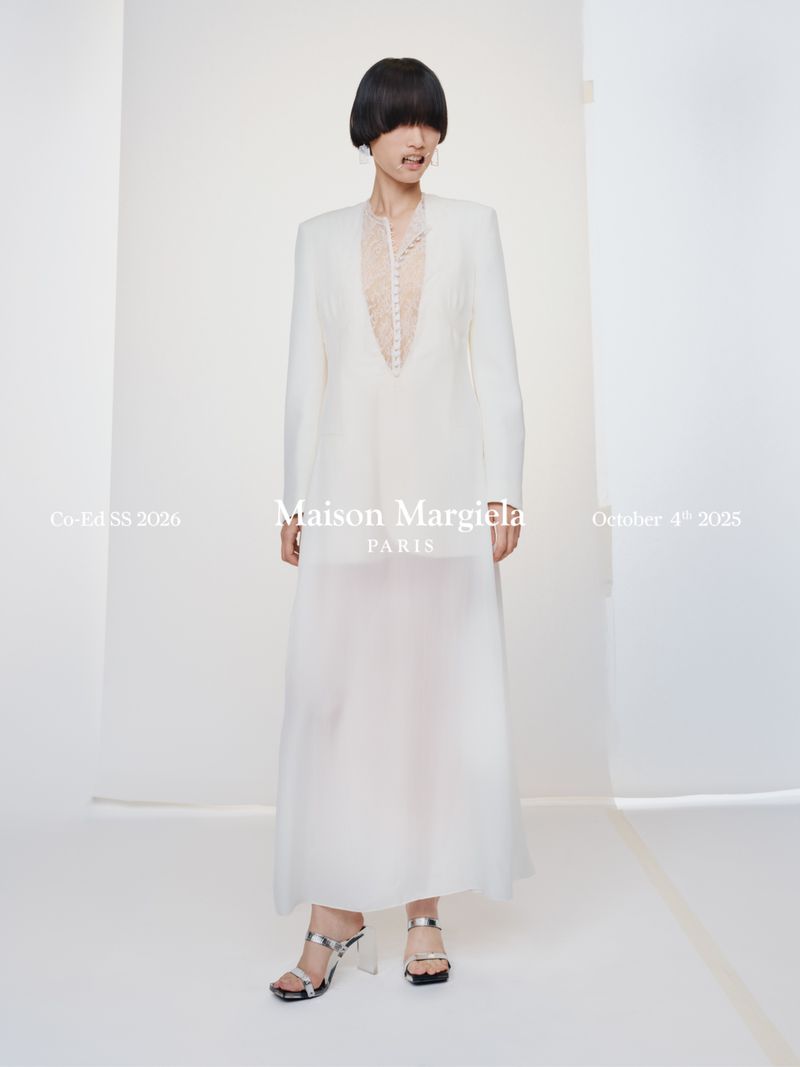 Runway shot of a model showcasing a Maison Margiela white dress with long sleeves and silver heels in a studio setting.