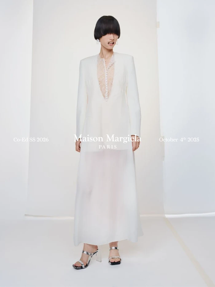 Model in a sheer white long dress and structured blazer standing against a white seamless background