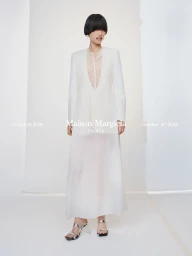 Model in a sheer white long dress and structured blazer standing against a white seamless background
