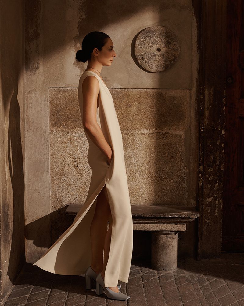 A slender woman with dark hair gazes in profile, wearing a long sleeveless cream dress and gray heels, set against a textured wall.