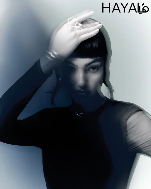 Portrait of a woman with hands near her head, wearing jewelry, styled in a black mesh top against a blended blue background.