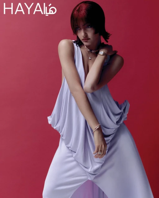 Fashion editorial shot of a woman with red-streaked hair wearing a lilac draped top and pants, posing against a red background indoors.