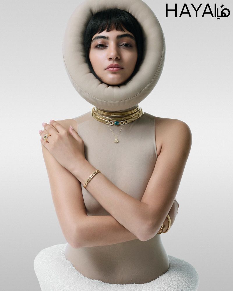 Close-up shot of a stylish young woman with a fringe hairstyle posing in a beige top and gold jewelry against a gray studio backdrop.