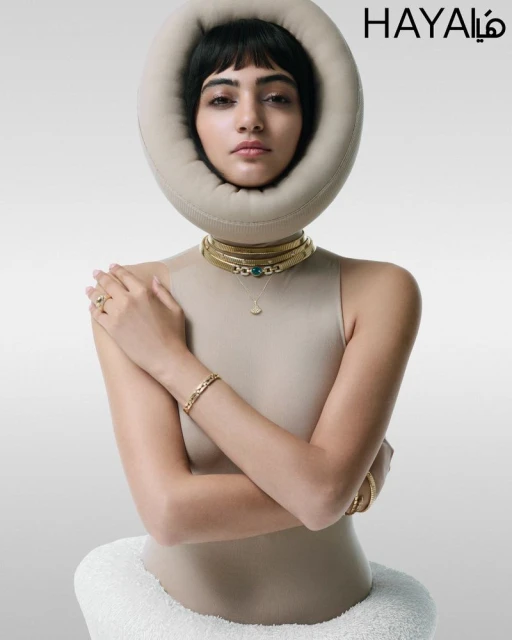 Close-up shot of a stylish young woman with a fringe hairstyle posing in a beige top and gold jewelry against a gray studio backdrop.