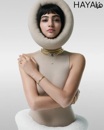 Close-up shot of a stylish young woman with a fringe hairstyle posing in a beige top and gold jewelry against a gray studio backdrop.