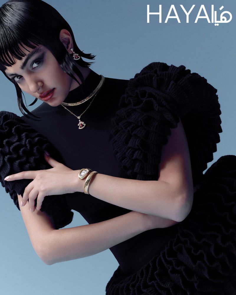 An adult woman in a black dress and jewelry poses in front of a light blue background for a beauty and fashion advertisement.