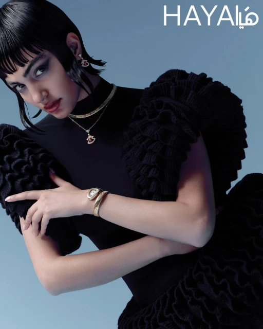An adult woman in a black dress and jewelry poses in front of a light blue background for a beauty and fashion advertisement.