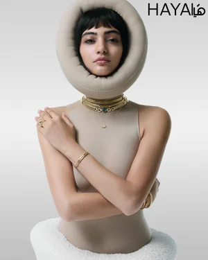 Model Soulin Omar framed by a round beige padded collar wearing layered gold jewelry against a studio background.
