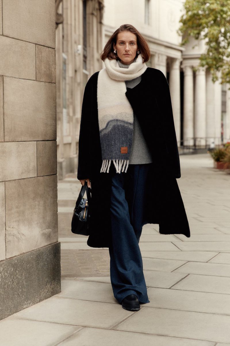Woman in a black coat and wide-leg jeans walking in an urban environment with a gray scarf and black purse.