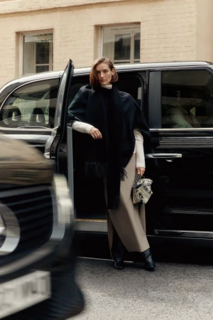 A stylish woman in a poncho stands by a black taxi, exuding fashion and urban sophistication in an outdoor setting.