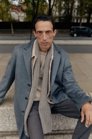 An adult man wearing a grey overcoat and scarf poses while sitting on a stone wall in a city with trees and a car in the background.