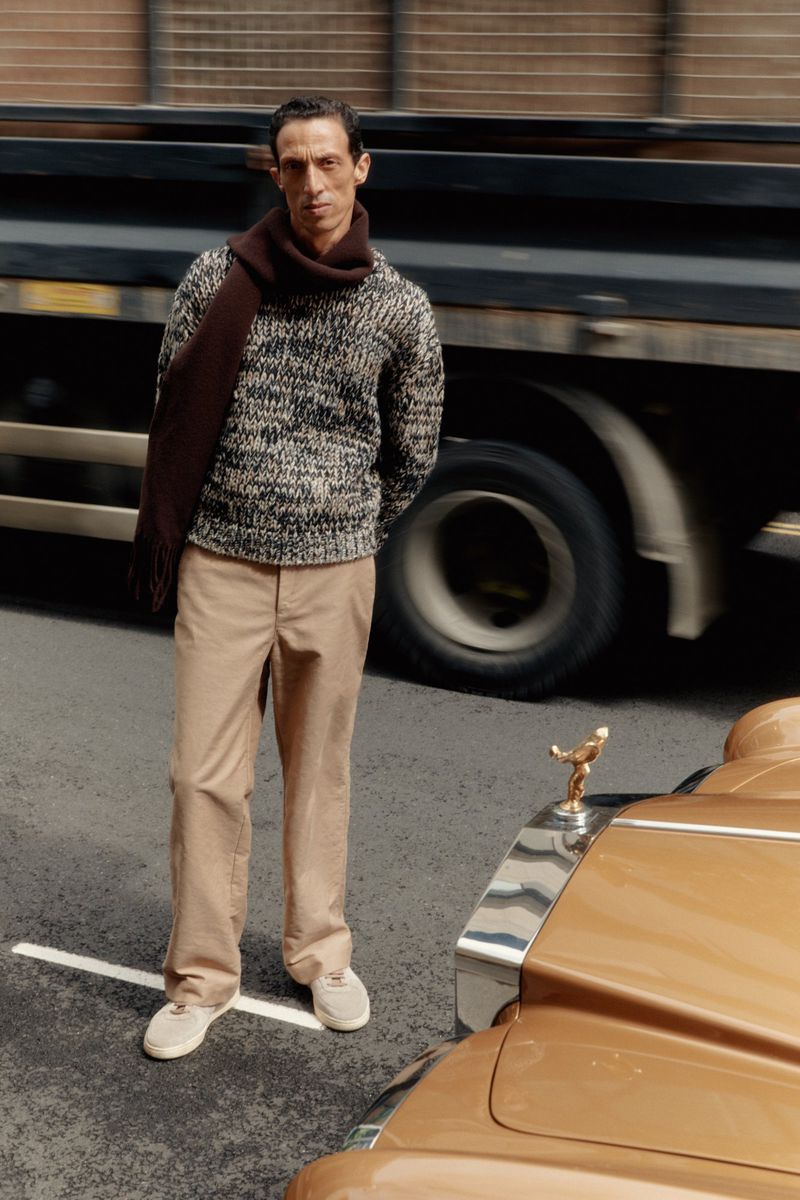 Mature adult male poses in an urban environment wearing a sweater, scarf, and tan pants next to a vintage tan Rolls Royce.