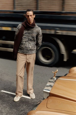 Mature adult male poses in an urban environment wearing a sweater, scarf, and tan pants next to a vintage tan Rolls Royce.