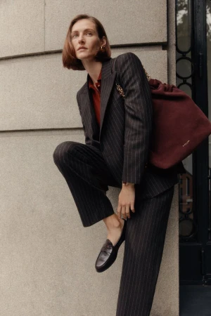 Elegant woman in pinstripe suit and loafers poses with maroon handbag against neutral wall, showcasing sophisticated fashion and style.
