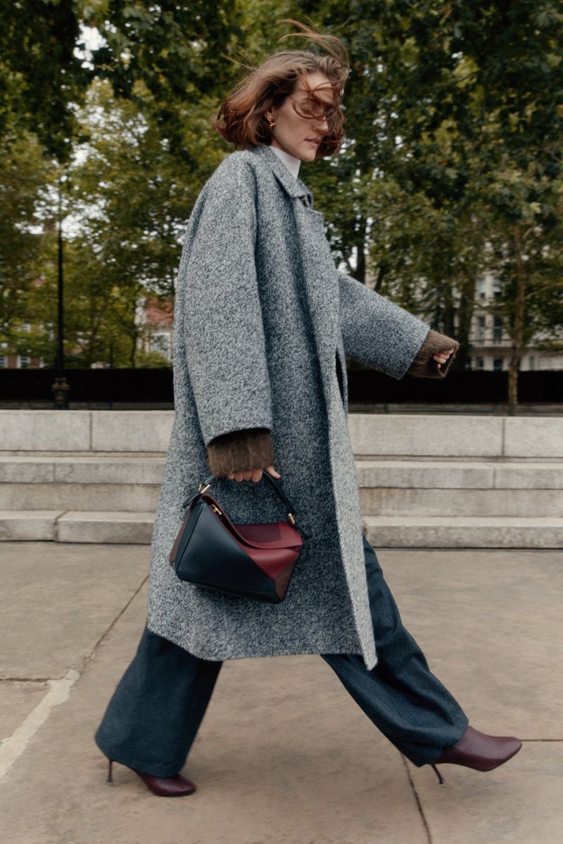 Woman in gray coat walks on concrete path, holding a handbag and wearing heeled boots, with trees and buildings in the background.