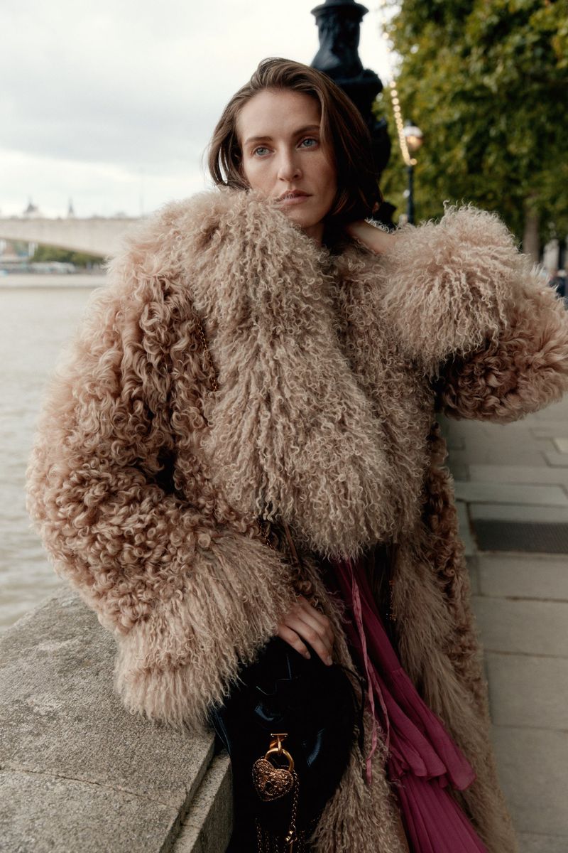 A woman with medium-length hair in a textured fur coat holds a small black purse with a gold medallion, with a river scene behind her.
