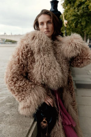 A woman with medium-length hair in a textured fur coat holds a small black purse with a gold medallion, with a river scene behind her.