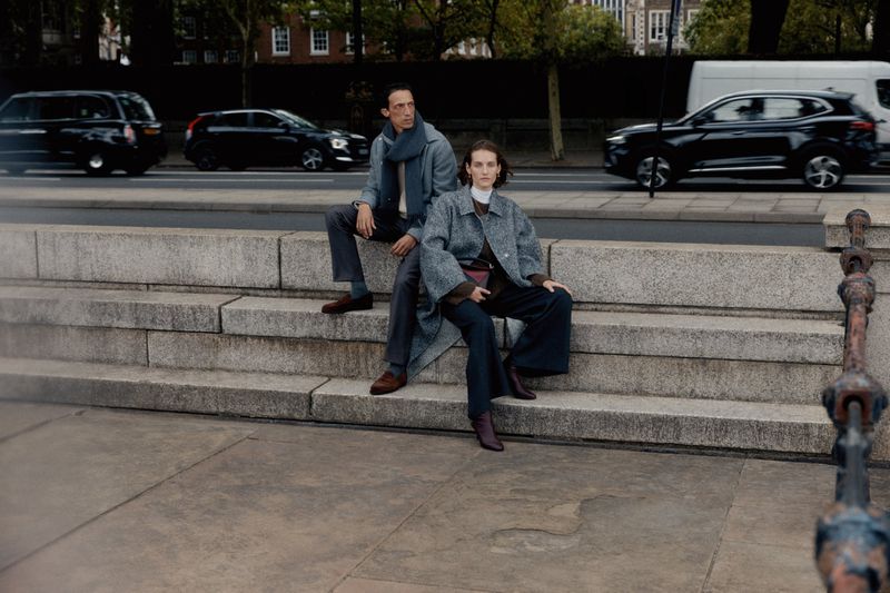 Two fashionably dressed people, a man and a woman, are sitting on stone steps. Cars and buildings line the street behind them.