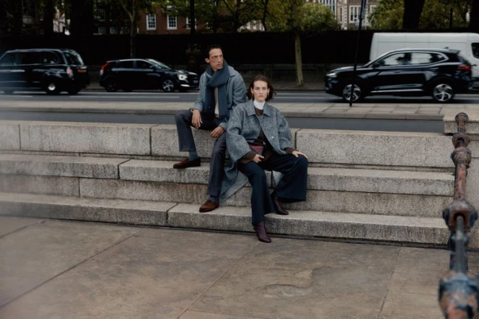 Two fashionably dressed people, a man and a woman, are sitting on stone steps. Cars and buildings line the street behind them.