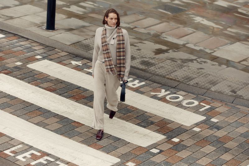 Adult woman crosses street on marked crosswalk in casual sweater, pants, scarf and loafers, holding purse with buildings in background.