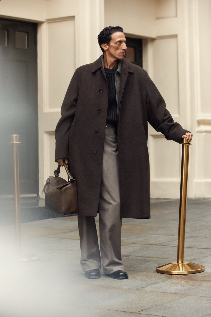 Stylish adult man in a dark coat and grey pants stands with a bag, accessorized with a gold stanchion in an outdoor setting.
