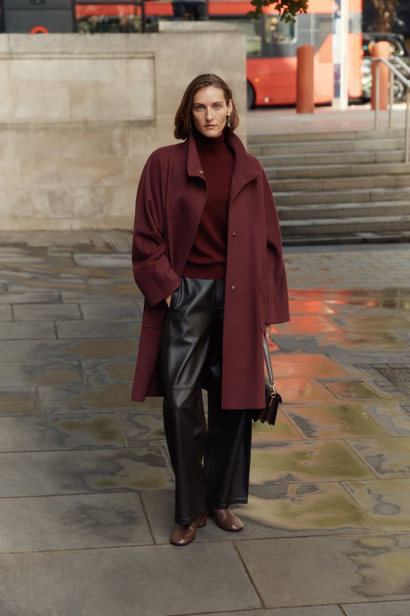 Woman in a longline maroon coat, turtleneck sweater, and faux leather pants walking in the city with a dark brown cross body purse.