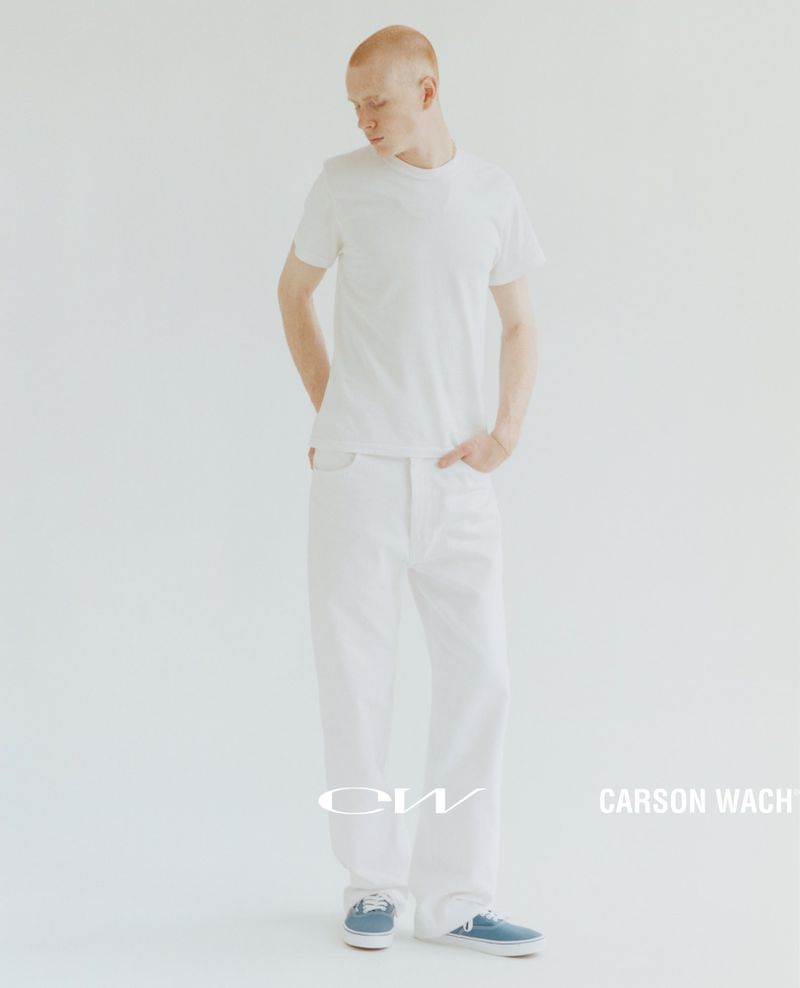 Full-length portrait of a male model with short hair in a white t-shirt, white denim jeans, and blue tennis shoes, set against a clean white backdrop.
