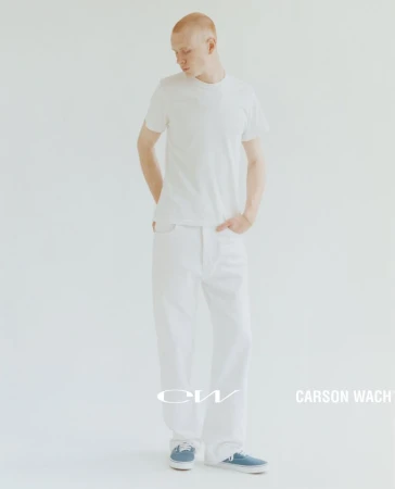 Full-length portrait of a male model with short hair in a white t-shirt, white denim jeans, and blue tennis shoes, set against a clean white backdrop.