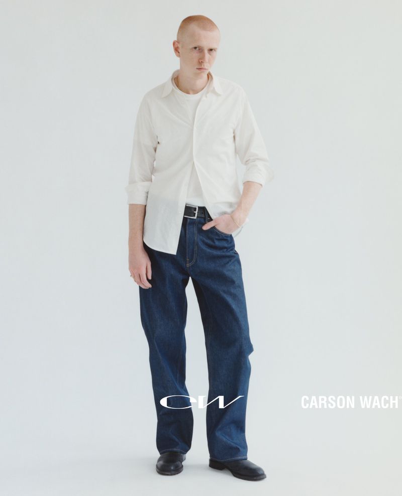 Full-length shot of a young adult man in a white shirt and wide-leg denim jeans, posing against a solid white backdrop in a studio setting.