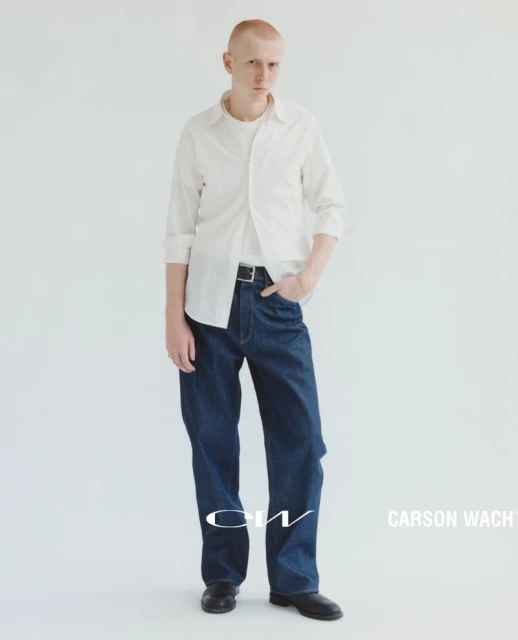 Full-length shot of a young adult man in a white shirt and wide-leg denim jeans, posing against a solid white backdrop in a studio setting.