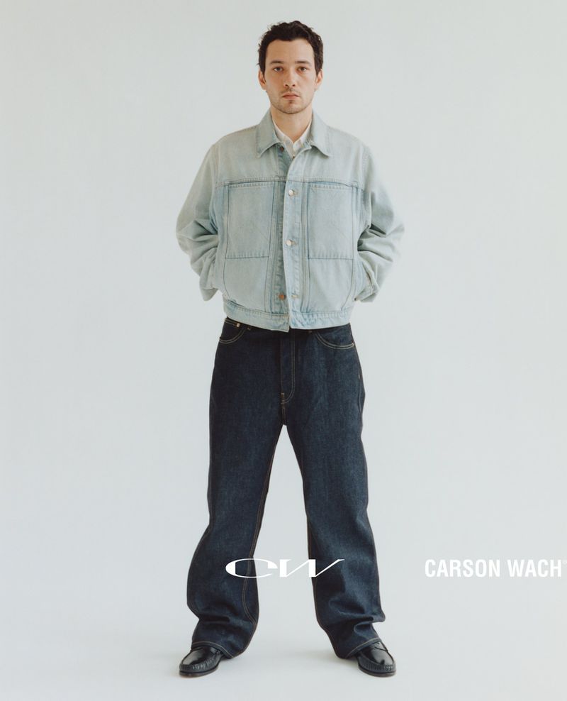 Full-length portrait of a man wearing a light denim jacket and dark blue jeans against a neutral studio backdrop