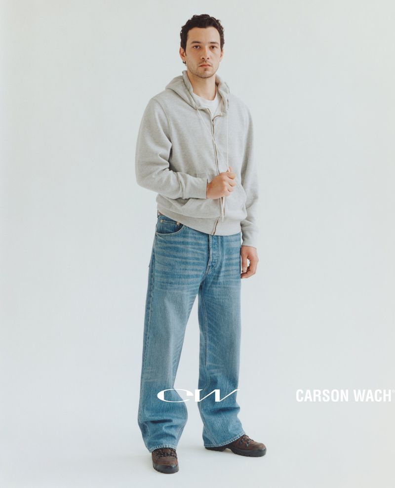 Full-length shot of a young adult male model wearing a grey hooded sweatshirt and wide-leg jeans, posing against a white backdrop.