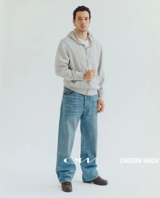 Full-length shot of a young adult male model wearing a grey hooded sweatshirt and wide-leg jeans, posing against a white backdrop.
