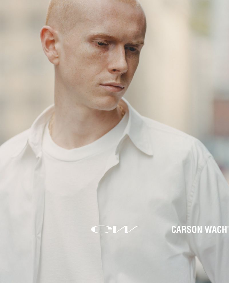 Portrait of a fair-skinned, bald adult male wearing a white button-down shirt against a blurred city background. The name Carson Wach is visible.