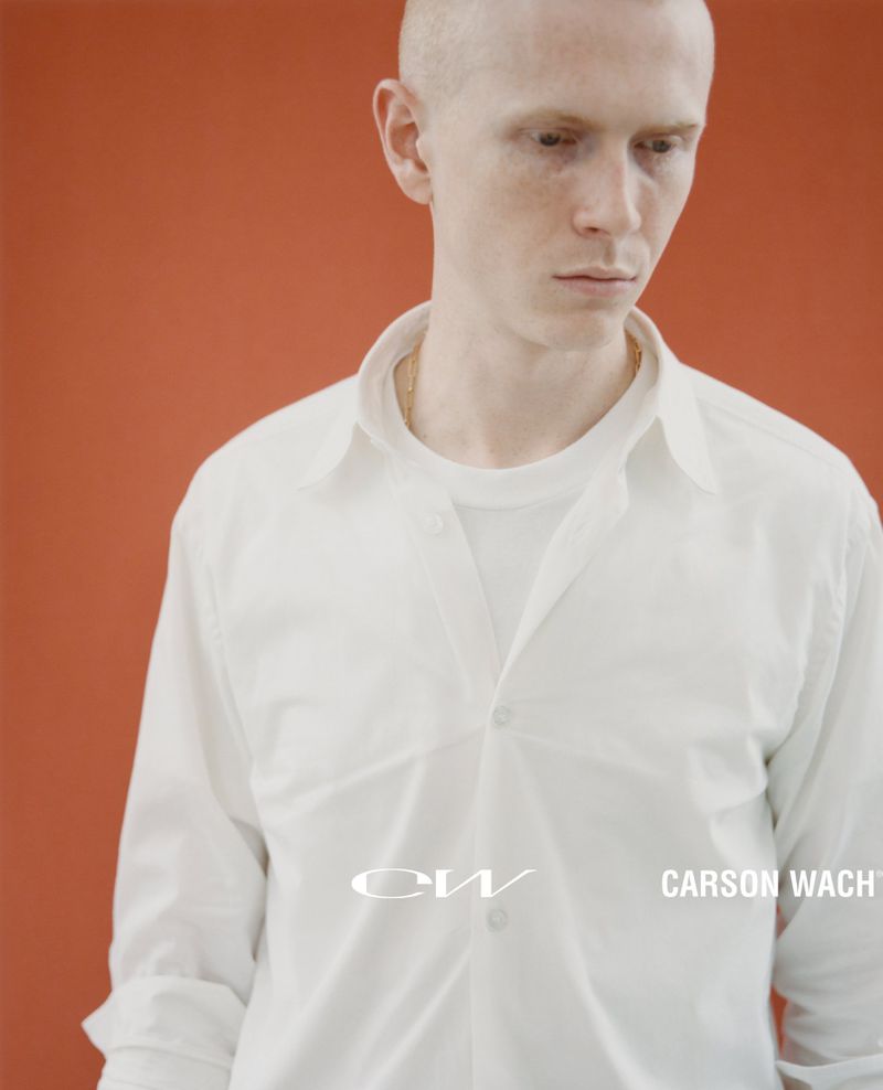 Portrait of a young adult with a shaved head wearing a white button-down shirt against an orange backdrop with text overlay.