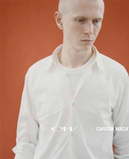 Portrait of a young adult with a shaved head wearing a white button-down shirt against an orange backdrop with text overlay.