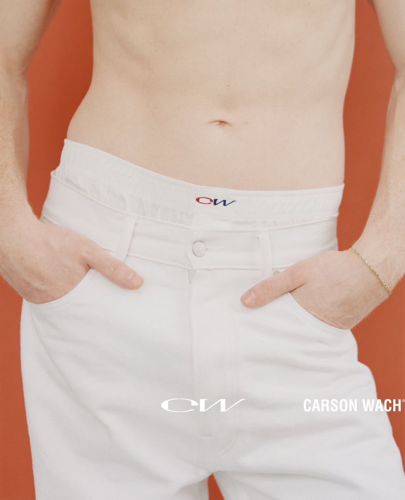 Torso of adult with hands in white denim jeans pockets and gold bracelet with a muted orange background and brand logos visible.