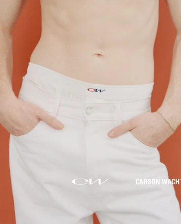 Torso of adult with hands in white denim jeans pockets and gold bracelet with a muted orange background and brand logos visible.