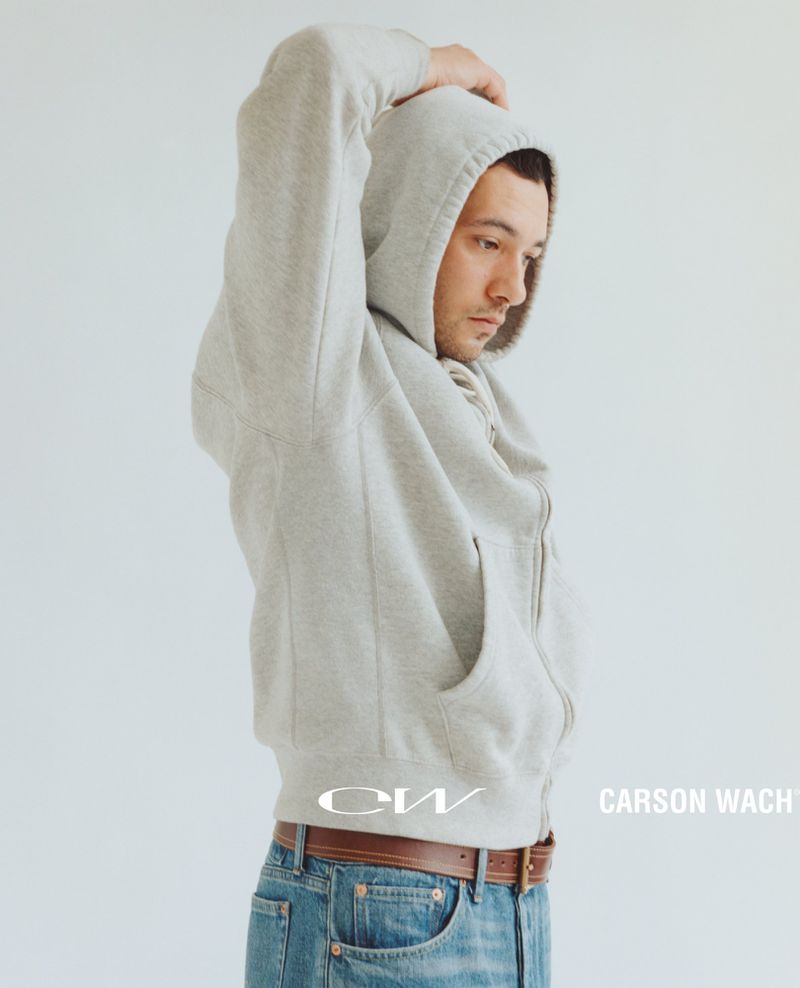 Man in a grey zip-up hoodie and blue jeans with one hand raised against a plain light background