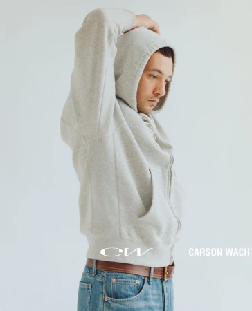 Man in a grey zip-up hoodie and blue jeans with one hand raised against a plain light background