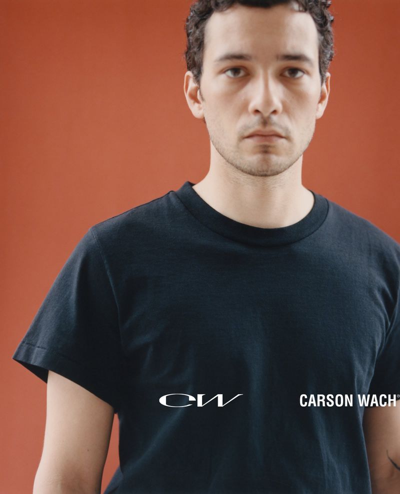 Portrait of a man with short curly dark hair wearing a black t-shirt against a rust-colored studio backdrop.