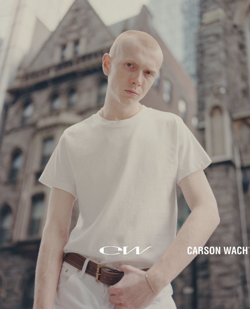 Bald man in a white t-shirt and white pants stands in an urban setting with large brick buildings and high rises in the background.