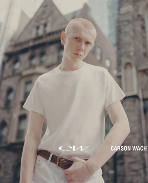 Bald man in a white t-shirt and white pants stands in an urban setting with large brick buildings and high rises in the background.