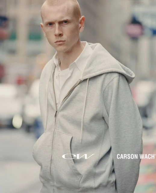Man in grey zip-up hoodie standing on city street. White collared shirt visible underneath. 'CW CARSON WACH' text on hoodie.