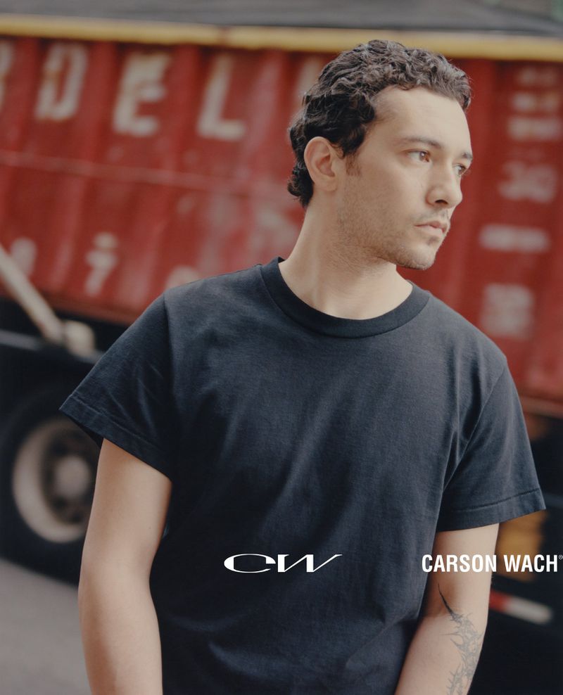 Portrait of a young adult man with curly dark hair in a black t-shirt outdoors with a blurry red truck in the background.