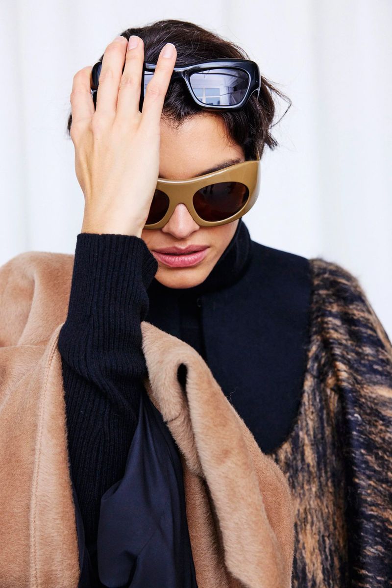 A woman with sunglasses on her face and on her head poses indoors, wearing a black top and a textured coat in a fashion setting.