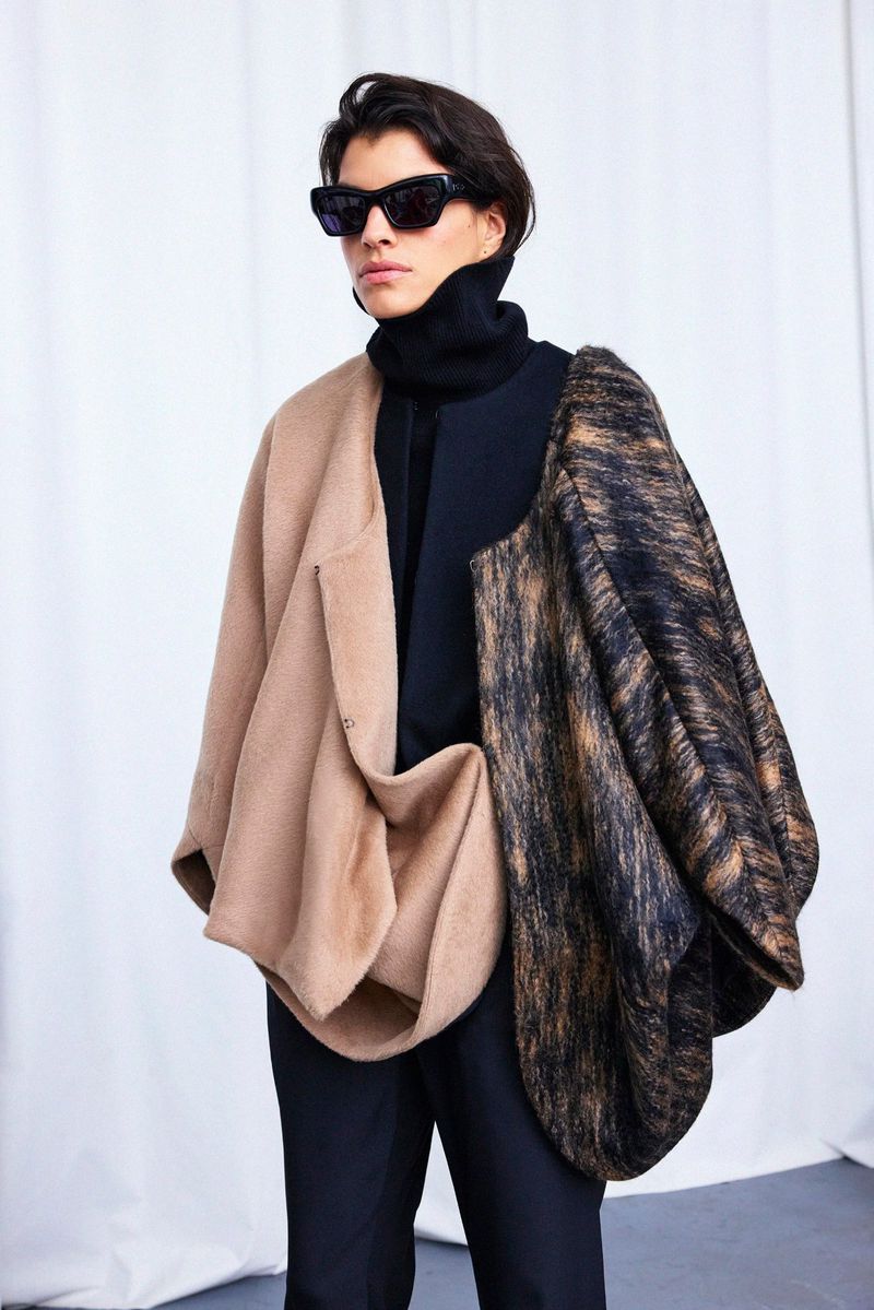 Fashionable individual wearing a unique beige and patterned poncho, stylish sunglasses, and a black turtleneck poses indoors against a white backdrop.