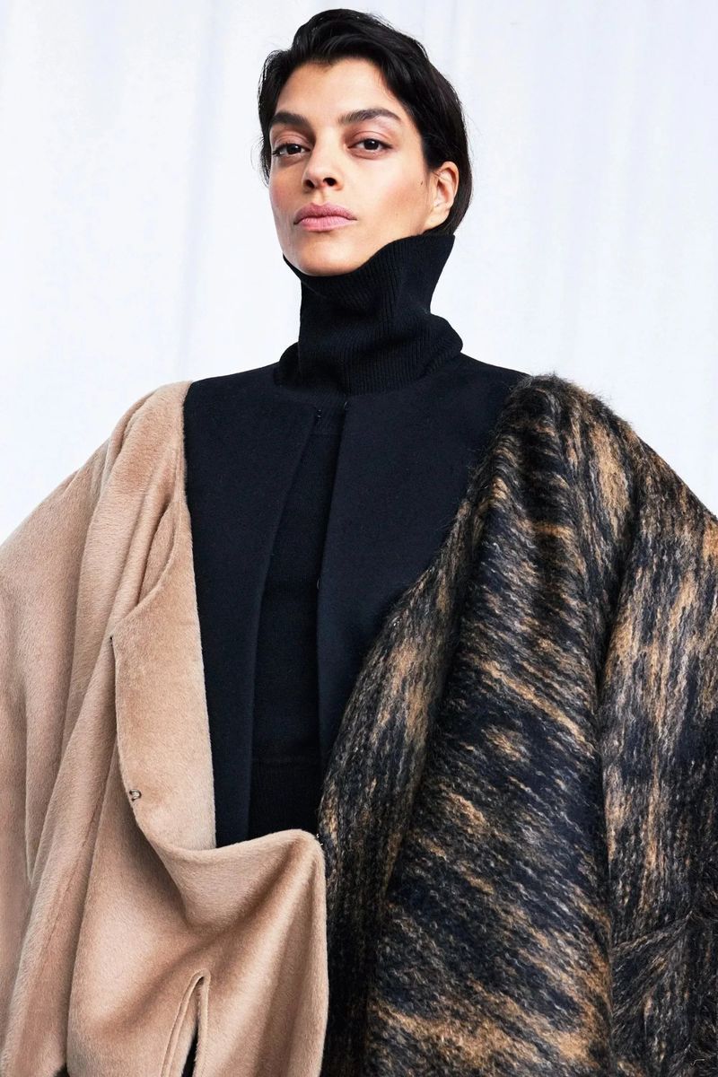 Fashionable woman poses in a black turtleneck, adorned with beige and printed brown overcoats, against a bright, neutral background.