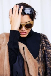 Model wearing sculptural tan visor sunglasses with black rectangular shades on her head against a plain white studio backdrop.